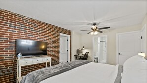 A hotel room with a brick accent wall, a flat-screen TV, a bed with white linens, a desk with a chair, and a ceiling fan. - 3BR Near Swamp Rabbit Trail • Fenced Yard • Pet-Friendly • 7 Min to Downtown (Greenville)