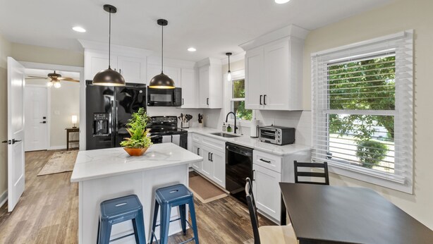 A modern kitchen with white cabinets, a central island with stools, a black refrigerator, a stove, and a sink. There is a window with blinds and a ceiling fan. - 3BR Near Swamp Rabbit Trail • Fenced Yard • Pet-Friendly • 7 Min to Downtown (Greenville)