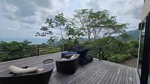 Eine Holzterrasse mit Sitzgelegenheiten im Freien, darunter eine Couch und Stühle, mit Blick auf eine Berglandschaft und das Meer.