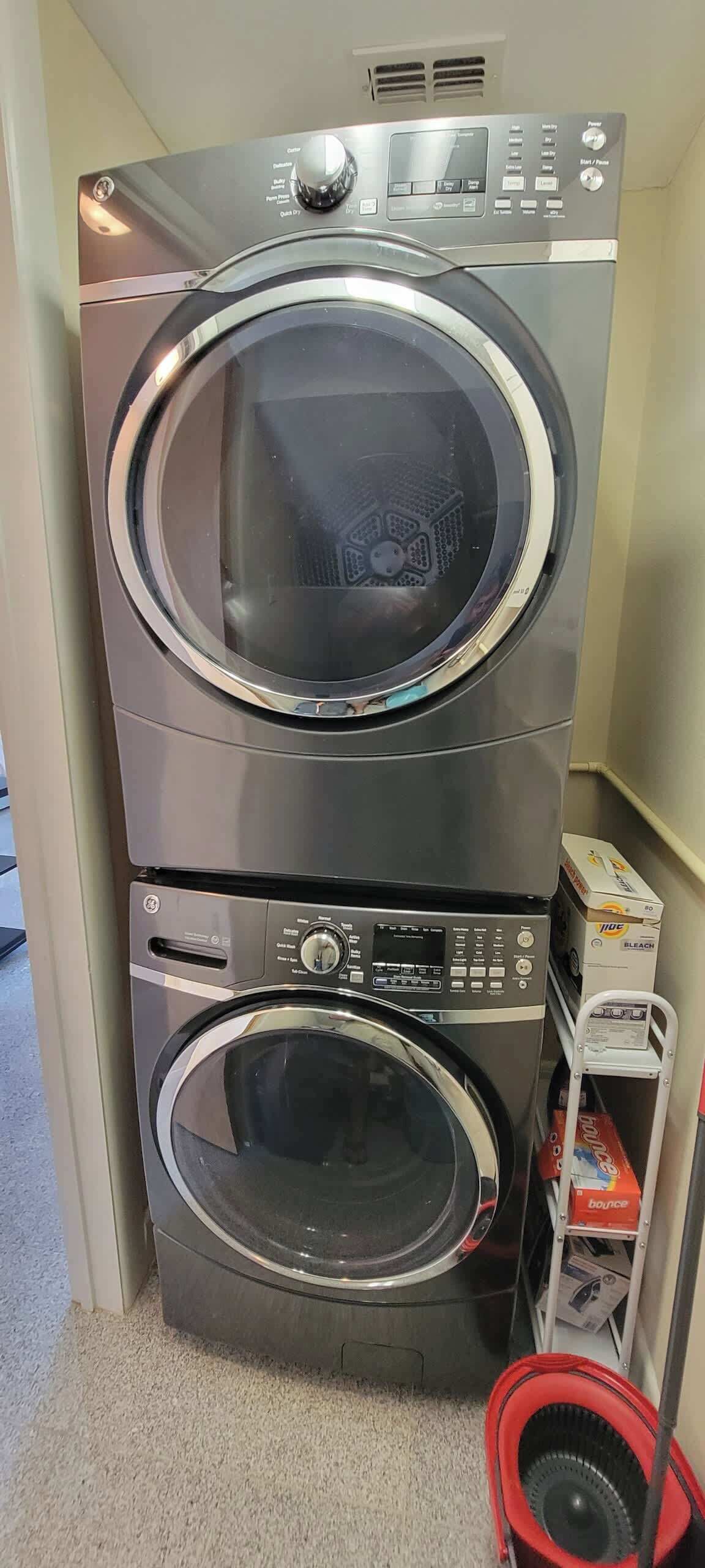 A stacked washer and dryer unit with control panels and a small shelf with detergent boxes.