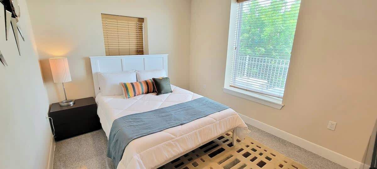 A bedroom with a bed, a nightstand, a lamp, a window with blinds, and a view of greenery outside.