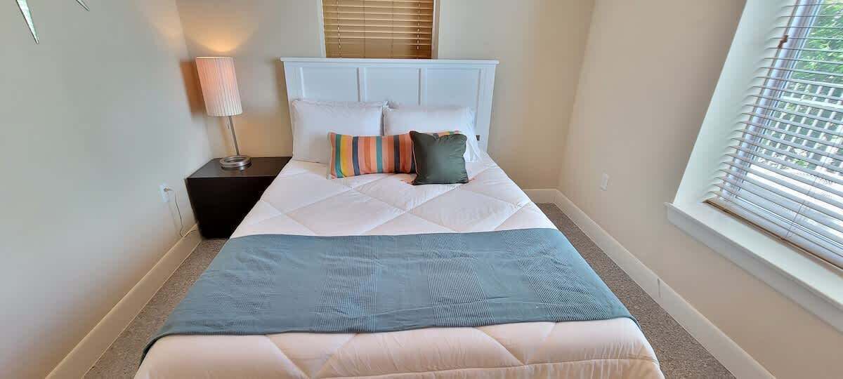 Una cama bien hecha con cabecera blanca, una lámpara de noche y una ventana con persianas.