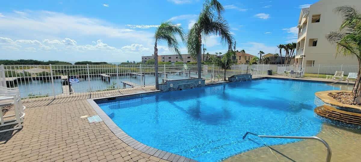 A swimming pool with a clear view of the water, surrounded by a brick walkway and palm trees.