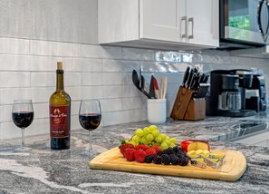 A kitchen counter with a bottle of wine, two glasses of wine, a cutting board with fruits and cheese, and a coffee maker.