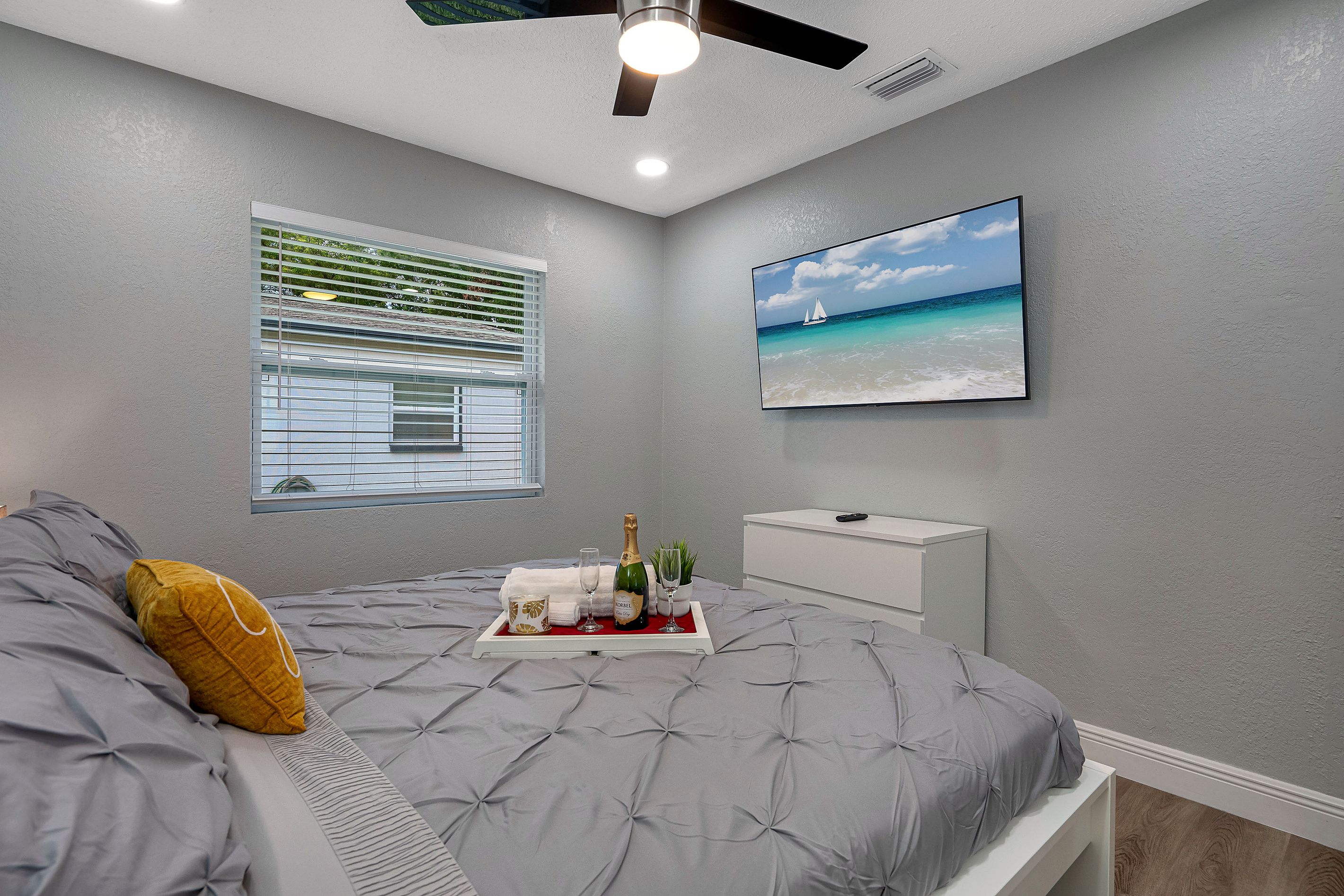 A bedroom with a bed, a window with blinds, a wall-mounted TV, and a framed picture of a beach scene.