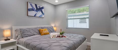 Quarto com cama, mesas de cabeceira, ventoinha no teto, janela com persianas e um quadro na parede.
