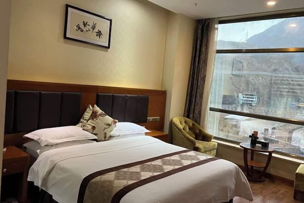 A hotel room with a large bed, a headboard, a chair, a small table, and a window with a view of a mountain and a cityscape.