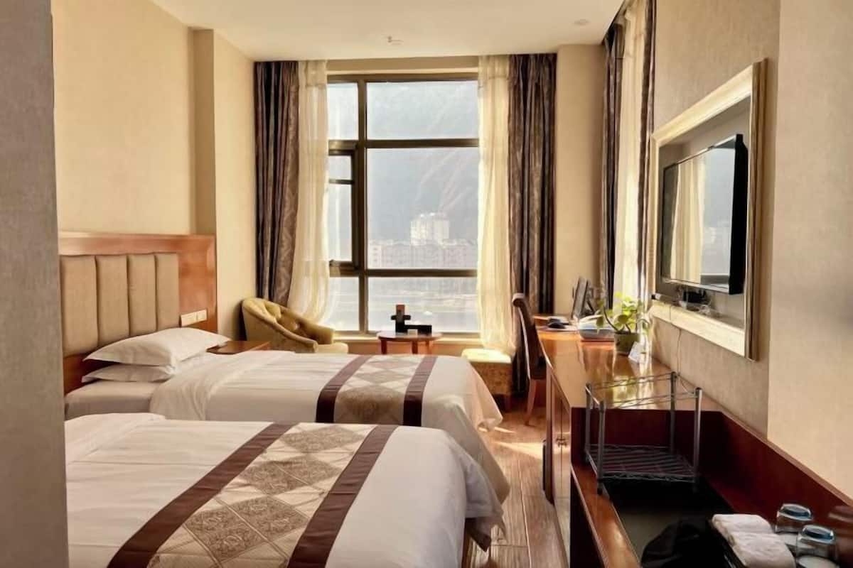 A hotel room with two beds, a desk, a chair, a TV, and a large window with a city view.