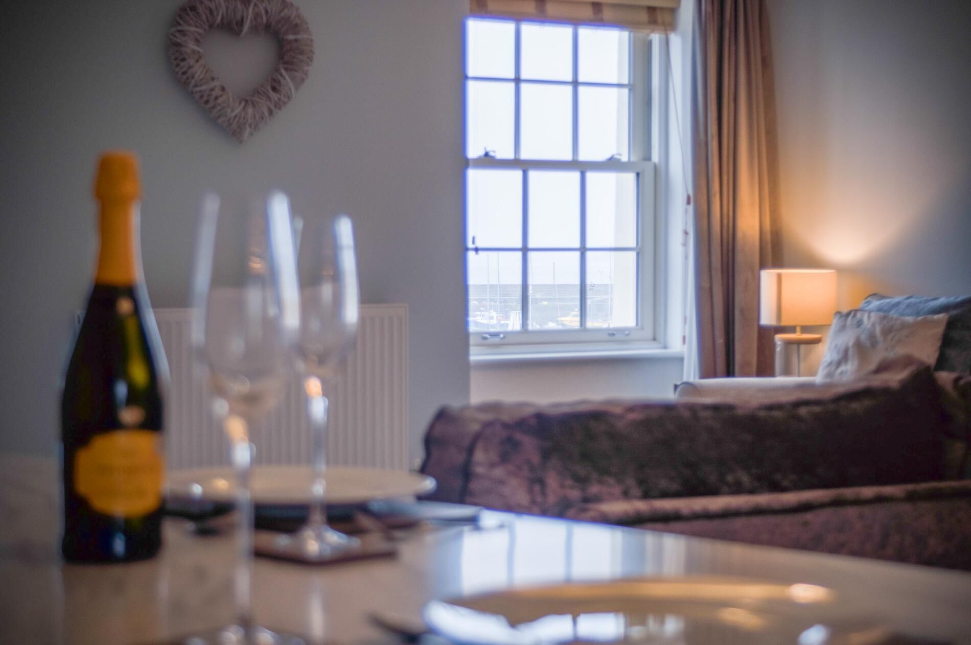 A room with a dining table set for two, a bottle of wine, and three wine glasses. There is a window with a view of the sea and a bed with a lamp in the corner.