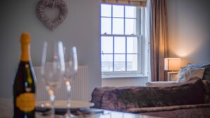 A room with a dining table set for two, a bottle of wine, and three wine glasses. There is a window with a view of the sea and a bed with a lamp in the corner.