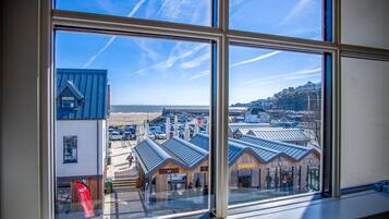 A view from inside a building with large windows overlooking a beach, a parking area, and a building with a sign that reads "intigo bee house".