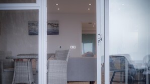 A room with a grey sofa, wicker chairs, and a table. There is a painting on the wall and a door leading to another room.