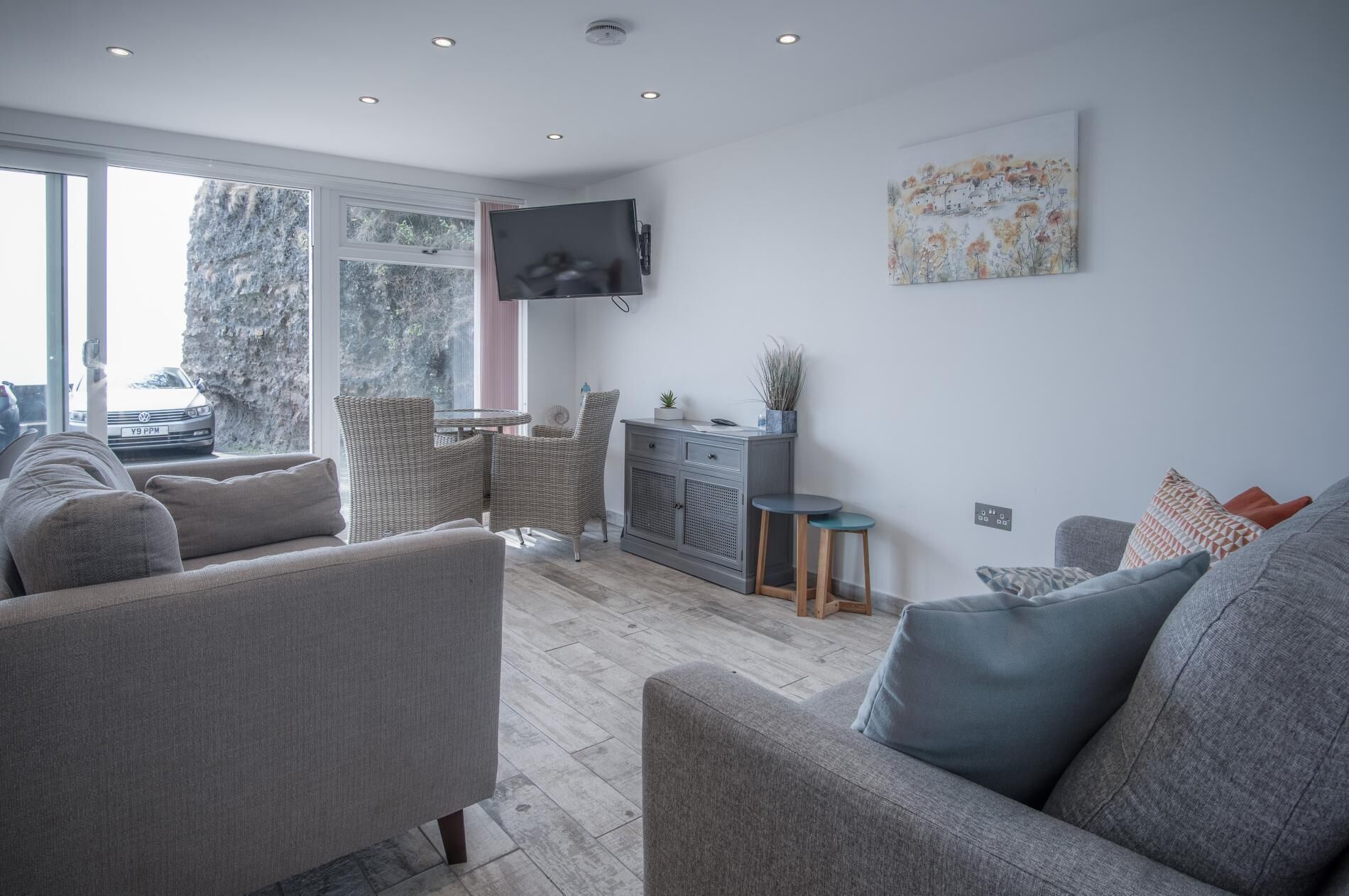 Un salon moderne avec des canapés gris, un coin repas avec des chaises en osier, un meuble gris et un téléviseur à écran plat fixé au mur.