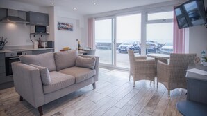A modern living room with a grey sofa, wicker chairs, a kitchen area with a microwave, and a large window overlooking parked cars.