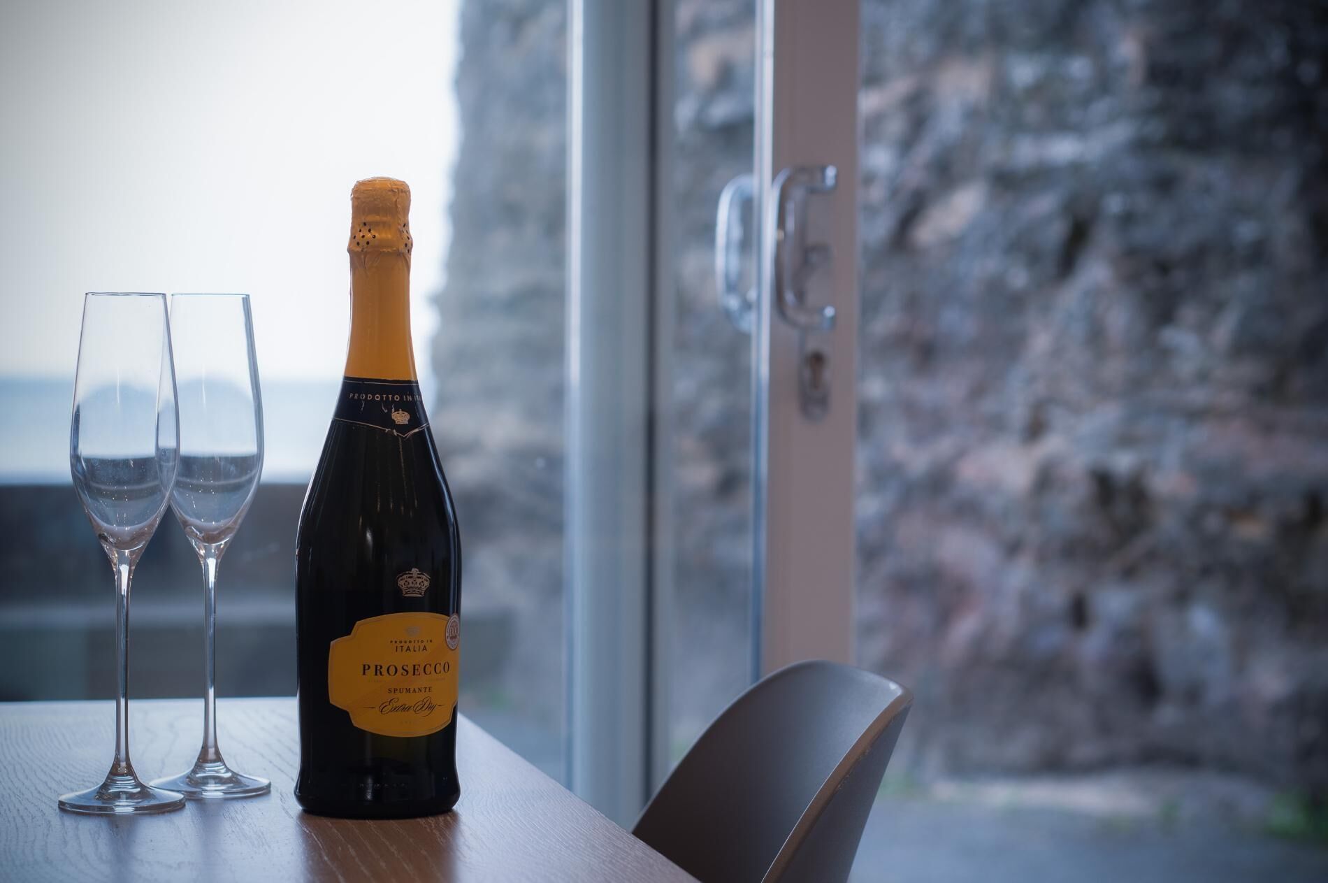 A bottle of Prosecco wine and two glasses on a table by a window with a view of a rocky landscape.