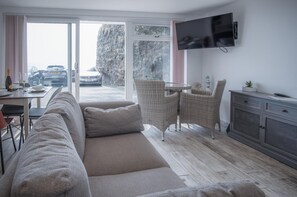 A modern living room with a sofa, a dining table, wicker chairs, a TV mounted on the wall, and a view of a car through the glass door.