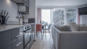 A modern kitchen with a dining area, large windows, and a grey sofa.