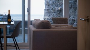 A modern living room with a sofa and a dining table, a bottle of wine, and a view of the sea through large windows.