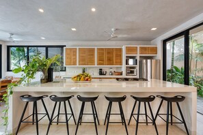 A modern kitchen with wooden cabinets, a stainless steel refrigerator, a fruit bowl, and a bar area with stools.