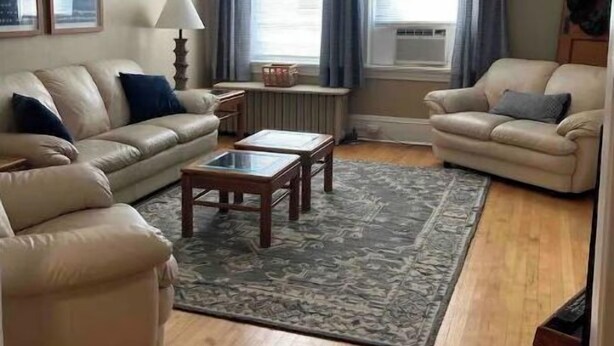 Sala de estar con dos sofás color beige, una mesa de centro de madera y un tapete estampado. Hay dos ventanas con cortinas, un ventilador de techo y cuadros enmarcados en la pared.