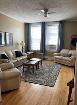 A living room with two beige sofas, a wooden coffee table, and a patterned rug. There are two windows with curtains, a ceiling fan, and framed pictures on the wall. - Downtown Living #1 (Fergus Falls)