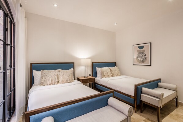 Quarto de hotel com duas camas, uma mesa de cabeceira, uma luminária, uma cadeira e um quadro na parede.
