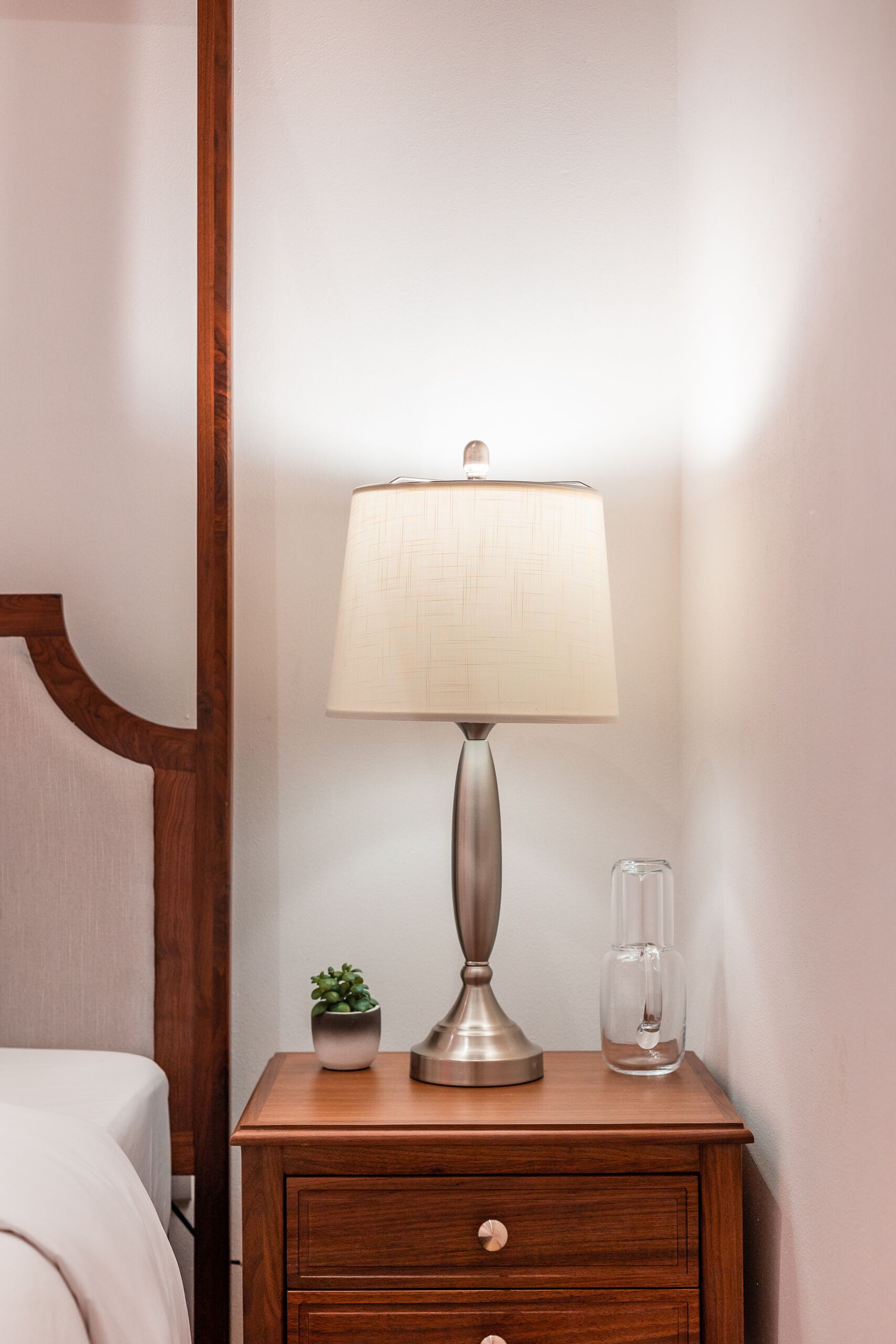 A wooden bedside table with a lamp, a small potted plant, and a glass container.