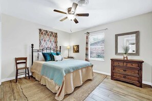 Une chambre à coucher comprenant un lit, une commode en bois, une chaise, un ventilateur de plafond, une fenêtre avec des stores et un miroir sur le mur.
