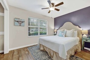 A bedroom with a bed, a ceiling fan, a window with blinds, a nightstand, and a patterned rug. - Billie Goat Haus-HotTub-Near Main St! (Fredericksburg)