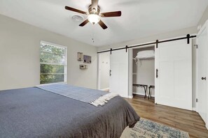 Une chambre à coucher comprenant un lit, un ventilateur de plafond, une fenêtre avec des stores, une porte donnant sur un placard et une petite table.