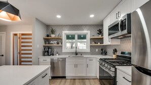 Eine moderne Küche mit weißen Schränken, einem Edelstahlkühlschrank, einem eingebauten Backofen und einem Gasherd. Ein Fenster bietet Blick ins Freie, und ein Regal beherbergt Küchenutensilien.