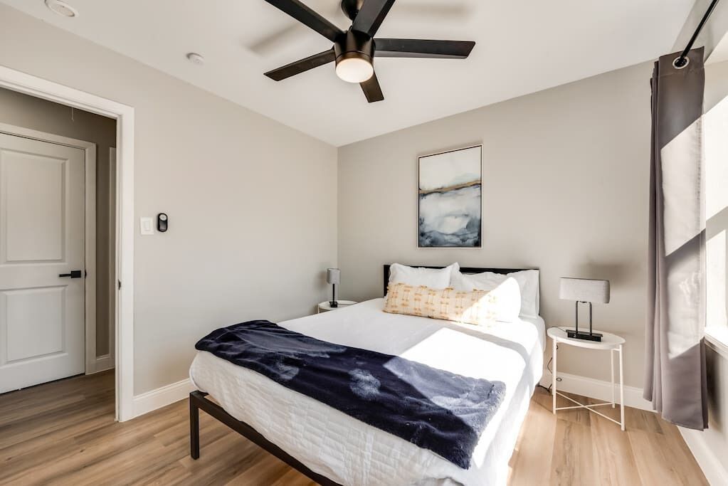 A bedroom with a bed, a ceiling fan, a nightstand, a lamp, and a painting on the wall.