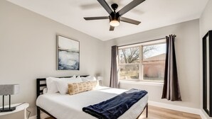 Ein Schlafzimmer mit Bett, Deckenventilator, Fenster mit Vorhängen, Nachttisch und Wandbild.