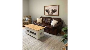 Een woonkamer met een bruine leren bank, een houten salontafel, een fauteuil met een patroon en een schilderij aan de muur.