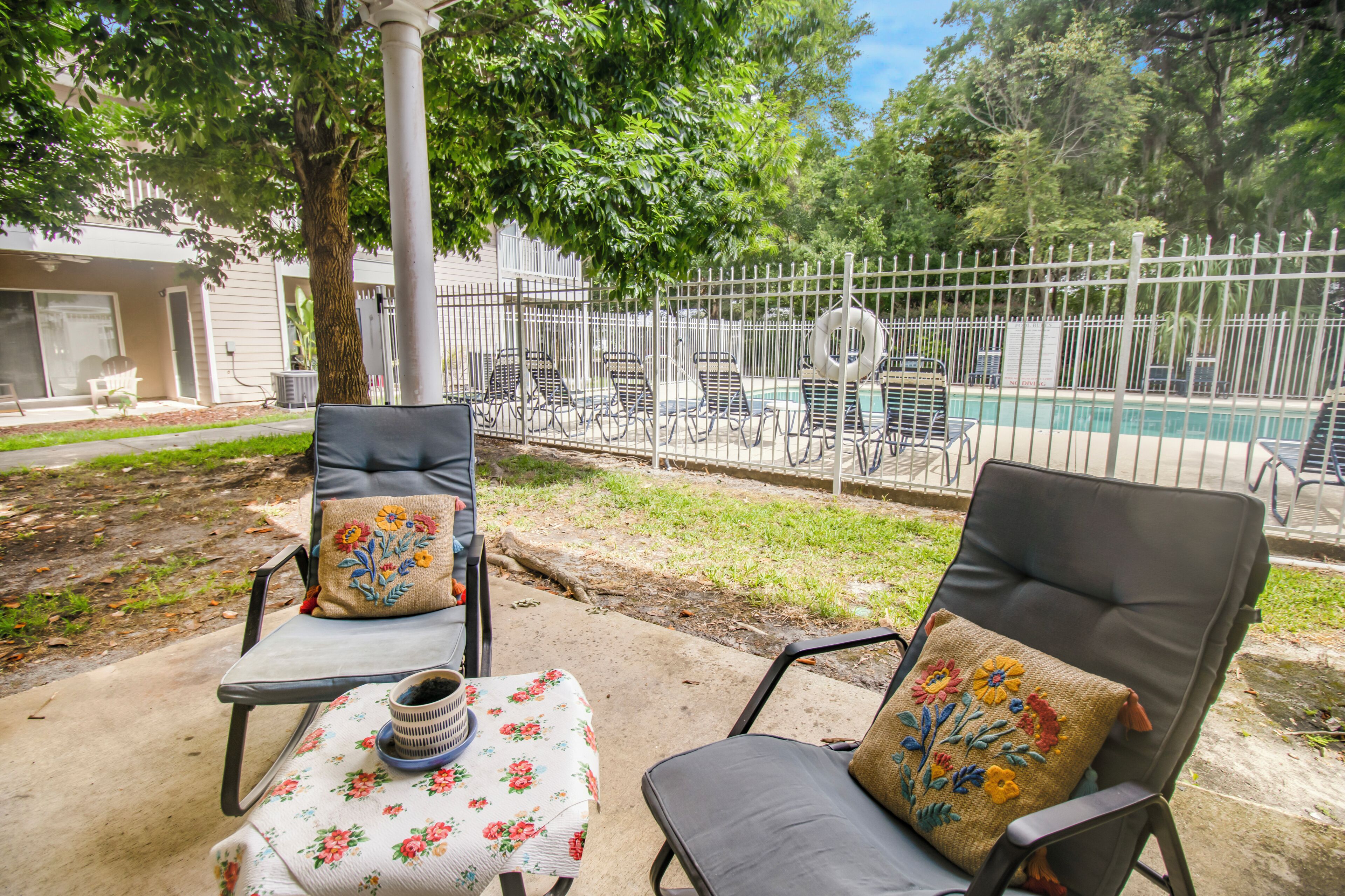 Deux fauteuils de jardin avec des coussins à motifs floraux, une petite table recouverte d’une nappe à motifs floraux, et une piscine en arrière-plan.