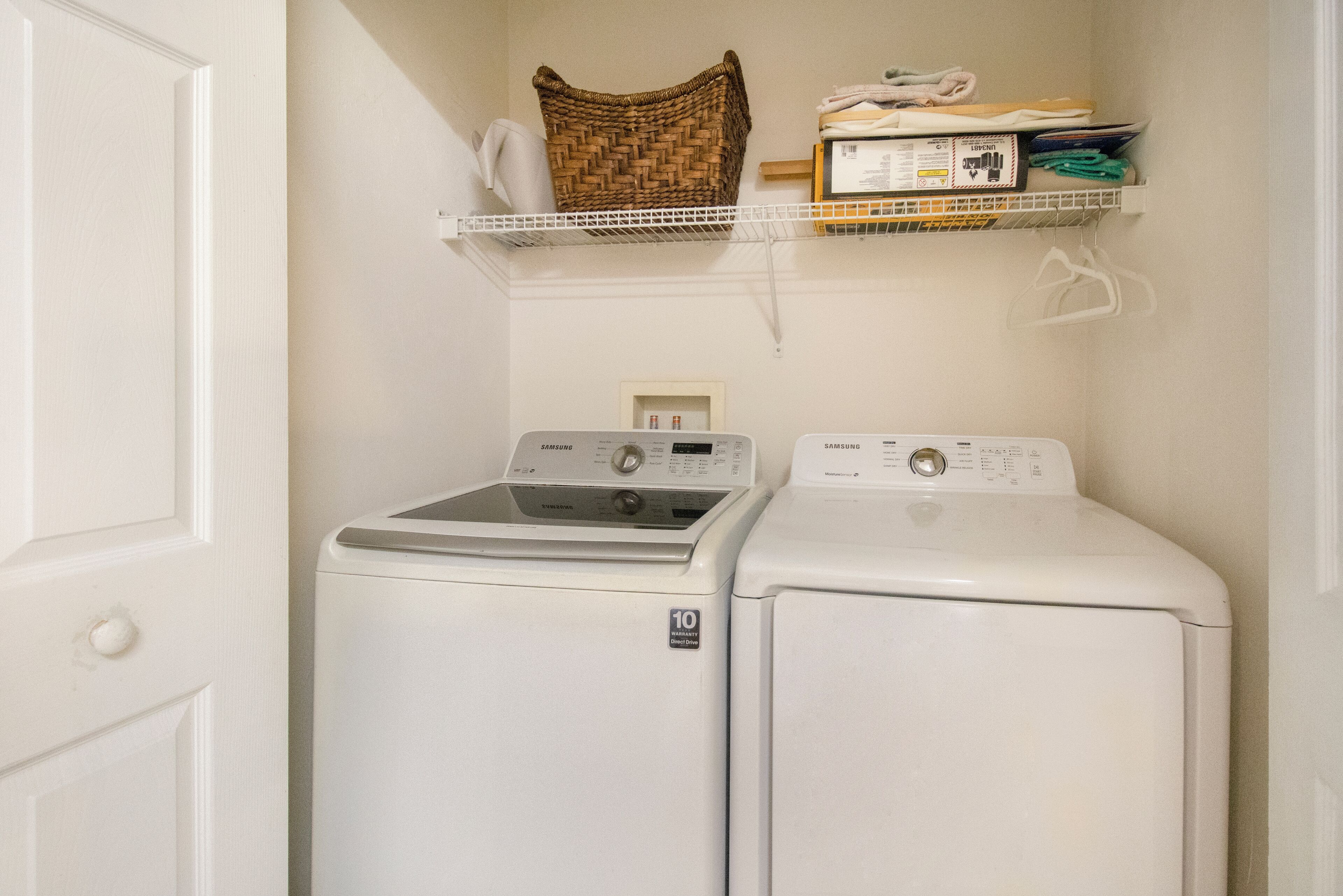 A laundry area with two white front-loading washing machines, a wicker basket, and a shelf with folded clothes and a box.