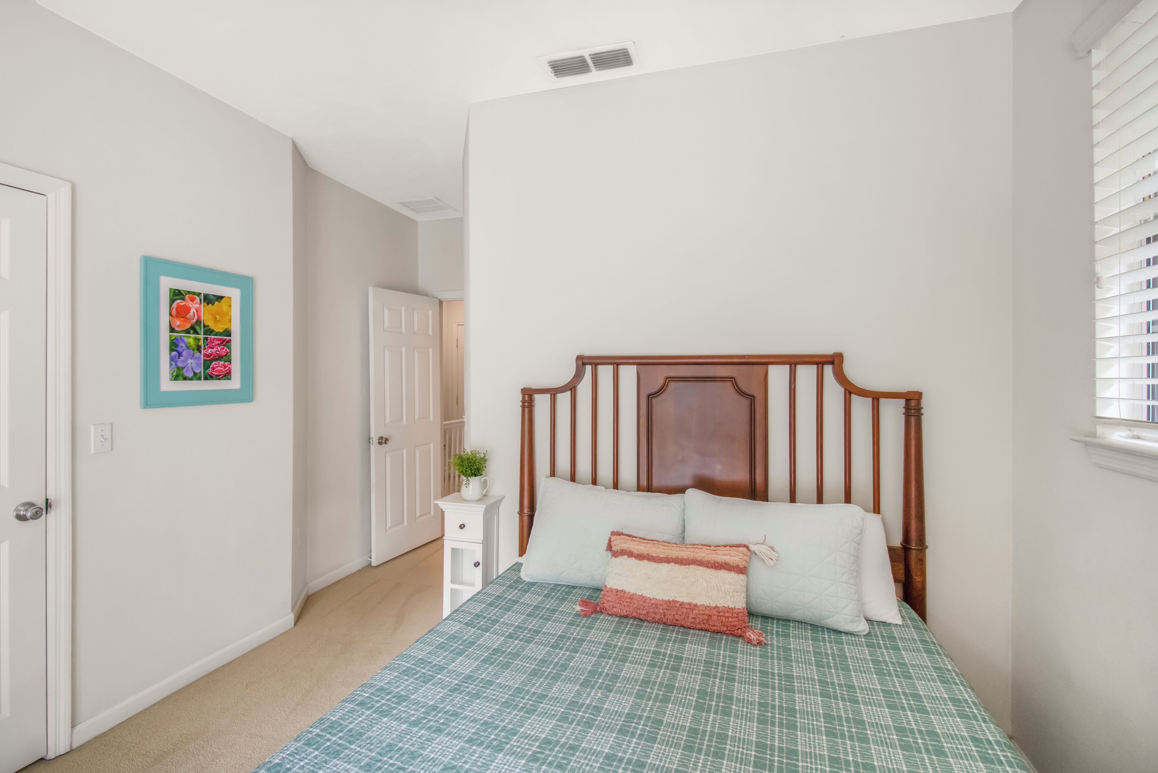 A bedroom with a wooden headboard, a green checkered bedspread, and a small white nightstand with a plant.