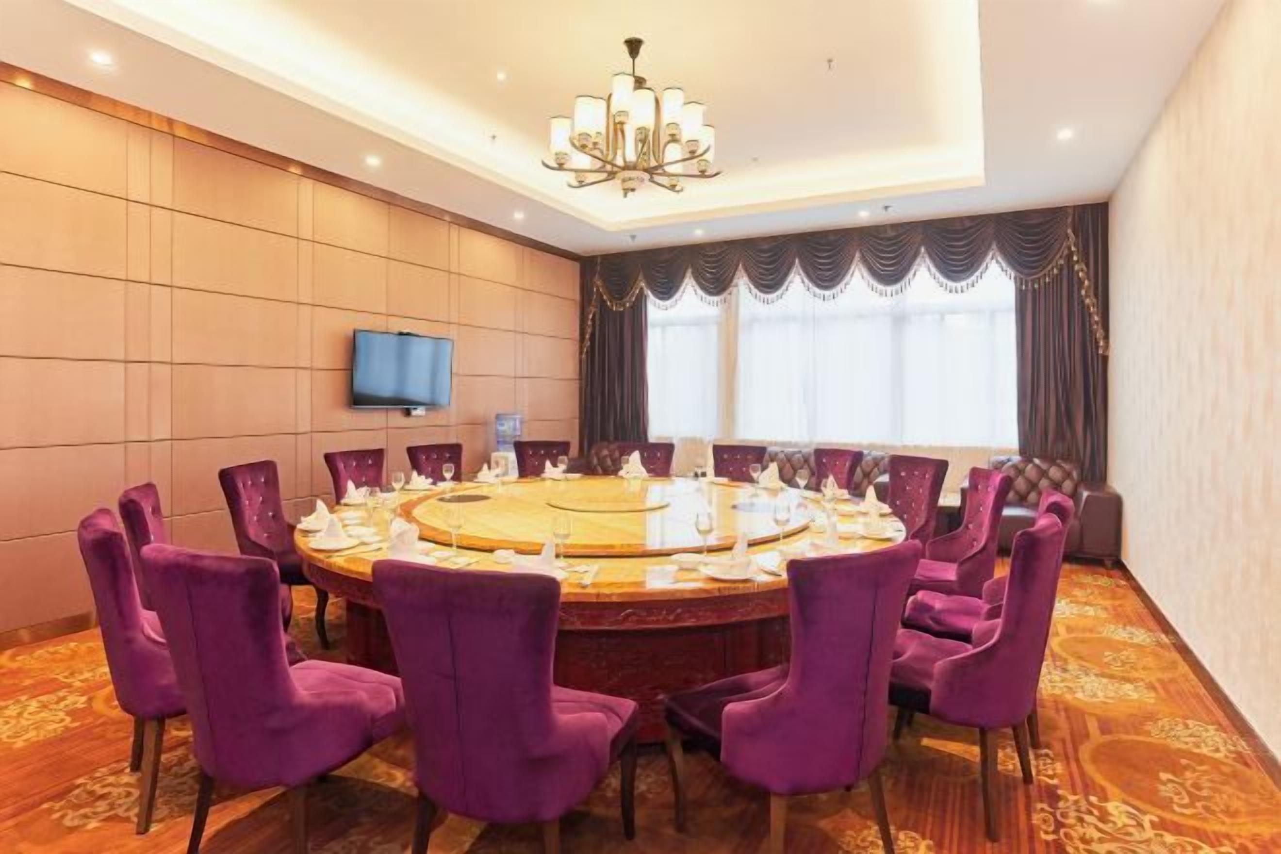 A conference room with a round table, purple chairs, a chandelier, a flat-screen TV, and a large window with curtains.