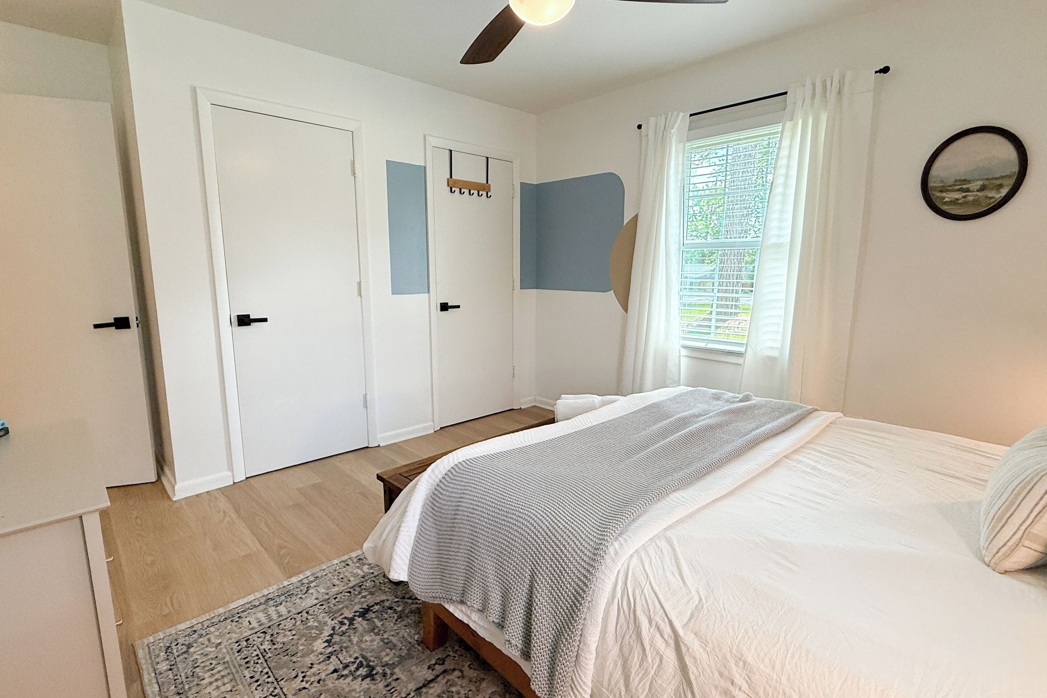 Ein Schlafzimmer mit Bett, einem fenster mit Vorhängen, einem Deckenventilator und einer Tür mit Kleiderhaken.