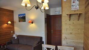 A cozy room with a wooden wall, a brown sofa, a wooden dining table, and two chairs. There are wall-mounted lamps and a poster on the wall. - 100m from ski slopes, Ground floor, balcony, 29m², Valloire (Valloire)