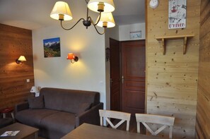 Um quarto aconchegante com parede de madeira, um sofá marrom, uma mesa de jantar de madeira e duas cadeiras. Há luminárias fixadas na parede e um cartaz.