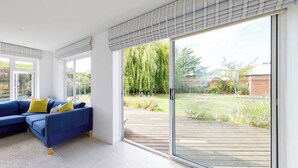 A modern living room with a blue sofa, a glass door leading to a wooden deck, and a view of a garden. - Secluded rural retreat, close to beach - sleeps 12 (West Sussex)