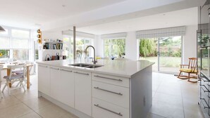 A modern kitchen with white cabinetry, a large island, and a built-in sink. There is a dining area with a table and chairs, and a view of the garden through large windows. - Secluded rural retreat, close to beach - sleeps 12 (West Sussex)