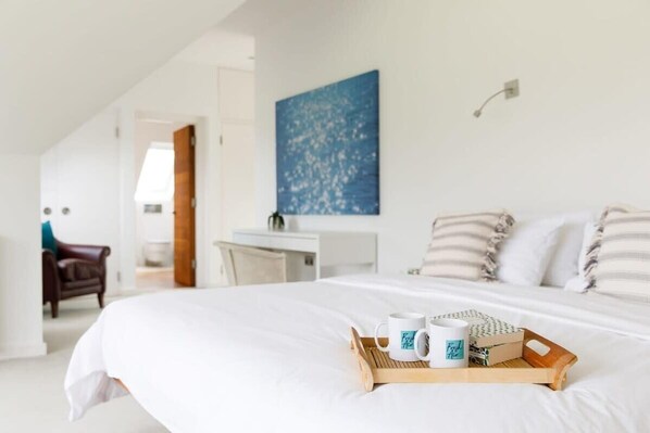 A neatly made bed with a tray of breakfast items, a blue abstract painting on the wall, and a view of a hallway with a door and a chair.