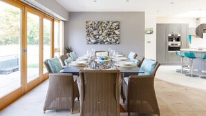 A dining area with a large table set for six, surrounded by wicker chairs, a stone wall art piece, and a view of the outdoors through large windows. - Goodwood Revival 6 Bed Retreat, Secure Parking (Birdham)