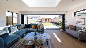 A modern living room with a glass coffee table, a sectional sofa, and a grey armchair. - Luxury designer coastal home for 10 with hot tub (West Wittering)