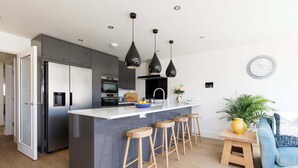 A modern kitchen with a central island, bar stools, a dining area with a table and chairs, and a clock on the wall. - Luxury designer coastal home for 10 with hot tub (West Wittering)