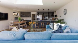 A modern living room with a blue sofa, a glass coffee table, and a kitchen in the background. - Luxury designer coastal home for 10 with hot tub (West Wittering)