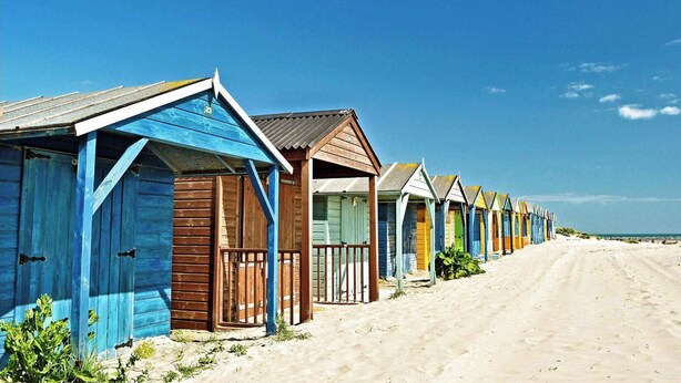 Une rangée de cabanes de plage, avec un sentier sablonneux menant à la plage.