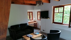 Ein gemütliches Wohnzimmer mit einer schwarzen Ledersofa, einem Holztisch und zwei schwarzen Sesseln. Ein Flachbildfernseher ist an der Wand montiert und es gibt ein Fenster mit Blick auf Grünflächen.